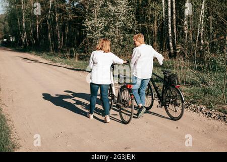 Zwei reife Freundinnen laufen mit Fahrrädern entlang des Waldes. Rückansicht Stockfoto