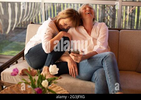 Zwei 55-jährige Damen in legerer Kleidung lachen fröhlich über Nachrichten, die sie auf dem Smartphone lesen Stockfoto