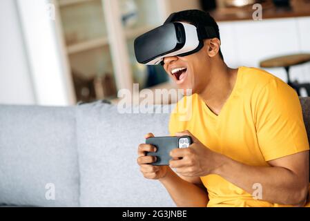 Aufgeregt Mixed Race Latino Kerl verwendet ein Virtual-Reality-Gerät, in einer digitalen Welt, hält Smartphone, spielen, während auf einem Sofa zu Hause in legerer Kleidung sitzen. Spielen in der virtuellen Welt Stockfoto
