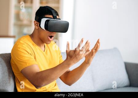 Moderner fassungslos hispanischer Kerl testet VR-Brille. Ein junger Mann nutzt Virtual-Reality-Brillen, blickt aufgeregt auf seine Hände, Gesten mit Händen, sitzt zu Hause auf dem Sofa im Wohnzimmer Stockfoto