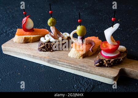Eine Auswahl an klassischen spanischen Tapas-Sandwiches mit Meeresfrüchten - Oktopus, Tintenfisch, Lachs, Wachtelei, Rettich, Grüner Salat und Pfeffer auf einem Brett auf einem schwarzen Rücken Stockfoto