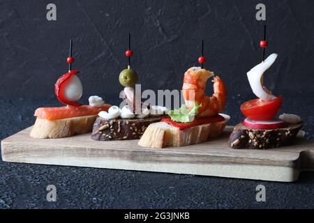 Eine Auswahl an traditionellen spanischen Tapas-Sandwiches mit Meeresfrüchten - Oktopus, Tintenfisch, Lachs, Wachtelei, Rettich, Grüner Salat und Pfeffer auf einem Brett auf einem schwarzen b Stockfoto