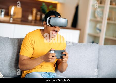 Glücklicher hispanischer junger Mann, der zu Hause in legerer Kleidung auf einem Sofa sitzt, eine VR-Brille verwendet, ein Virtual-Reality-Gerät, hält in einer digitalen Welt Smartphone und spielt in der virtuellen Welt Stockfoto