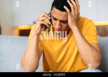 Unglücklicher, trauriger hispanischer junger Mann, der zu Hause auf einem Sofa sitzt, in legeres gelbes T-Shirt gekleidet, verwirrt, auf dem Smartphone redet, verwirrt, unangenehme Telefongespräche führt Stockfoto