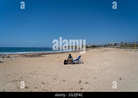 Trauriger Maskenmensch sitzt auf einer Sonnenliege inmitten eines leeren Strandes am Mittelmeer in Zypern. Leerer Strand, keine Touristen. Sperren und Reisen Stockfoto