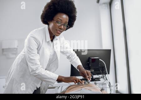 Nahaufnahme Porträt der jungen angenehmen afroamerikanischen Ärztin gp Kardiologe, kleben EKG-Elektroden für die Herzdiagnostik für junge Mann Patienten. Medizinische Geräte für Elektrokardiogramm Stockfoto