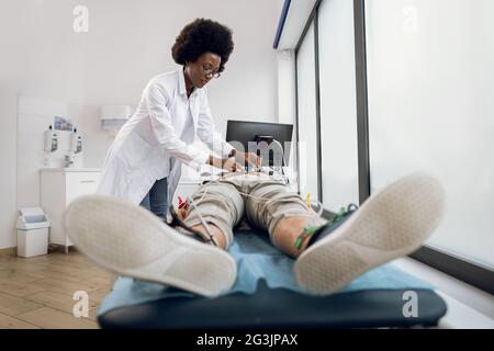 Herz-Elektrokardiogramm für jungen Mann Patienten. Der junge afroamerikanische Arzt legt Vakuumsensoren an den männlichen Patienten an, um sein Elektrokardiogramm in einer modernen Lichtklinik aufzunehmen Stockfoto