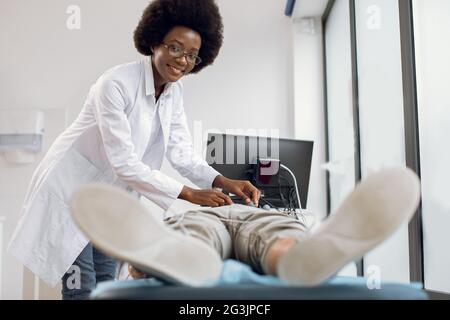 Medizin, Kardiologie, EKG des Herzens Konzept. Lächelnde, sympathische, hochqualifizierte afrikanische Ärztin legt Elektroden zur Elektrokardiographie an der Brust des männlichen Patienten an Stockfoto