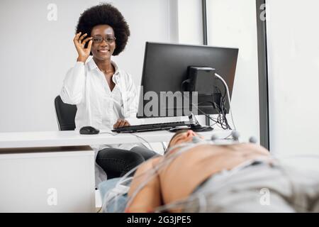 Prävention von Herz-Kreislauf-Erkrankungen. Fröhliche weibliche afrikanische Ärztin, sitzend am Tisch mit Monitor, EKG-Leitung. Männlicher Patient mit EKG-Elektrokardiogramm im Krankenhaus Stockfoto