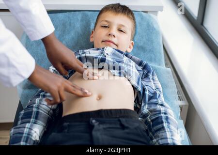 Netter lächelnder Junge im Alter von 10 Jahren, der auf der Couch in der modernen Klinik liegt und seinen Arzt besucht. Weibliche Afro-Kinderärztin, die den Abdomen eines Kindes mit manueller Palpation untersucht Stockfoto