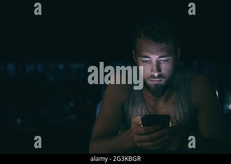 Dunkler Nachtmann, der auf den Handybildschirm schaut, der spät in der Nacht in Schlaflosigkeit im Bett oder draußen in der Stadt mit Skyline-Hintergrund SMS schreibt. Seriös aussehend Stockfoto