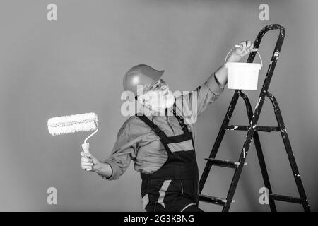 Arbeiten in der Werkstatt. Senior Mann Maler verwenden Rolle auf der Leiter. Malerei der Wand in rosa. Professioneller Maler in Arbeitskleidung. Arbeiter Malerei Stockfoto