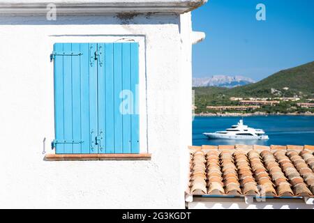 (Selektiver Fokus) atemberaubende Aussicht auf ein geschlossenes blaues Fenster an einer weißen Wand und eine unscharf getaufte Luxusyacht, die im Hintergrund segelt. Stockfoto