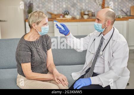 Ein Arzt misst die Temperatur einer älteren Frau in ihrem Zuhause mit einer Schutzmaske. Coronovirus-Infektionsdiagnosekonzept Stockfoto