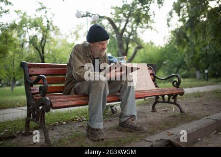 Obdachloser sitzt auf der Bank Holding Buch in den Händen. Stockfoto