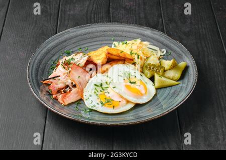 Spiegeleier, Schinken mit Kartoffelkeilen, eingelegter Kohl und Gurke auf schwarzem Holztisch Stockfoto