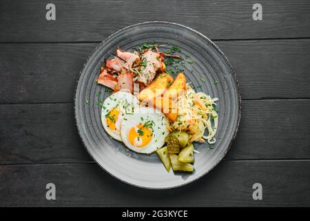 Blick von oben auf Spiegeleier, Schinken mit Kartoffelkeilen, eingelegtem Kohl und Gurke auf schwarzem Holztisch Stockfoto