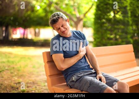 Mann mit Herzschmerzen draußen. Männlicher Brustschmerz, der auf der Bank im Park sitzt. Gesundheits- und Medizinkonzept Stockfoto