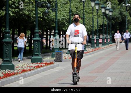 Kerl in schützende Gesichtsmaske reitet einen Elektroroller in Alexander Garten. E-Scooter fahren in der Sommerstadt Stockfoto