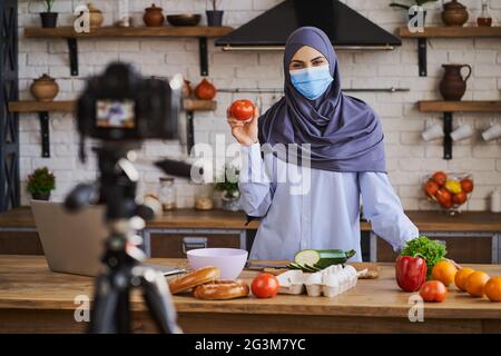 Fröhliche muslimische Frau, die eine Tomate in der Hand hält und mit der Kamera spricht Stockfoto