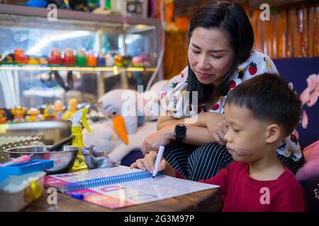 Junge Schüler Hausaufgaben zu Hause mit Schule Bücher, Zeitung und digitale Pad von seiner Mutter geholfen. Mom schreiben auf dem copybook Lehre seinen Sohn. Stockfoto