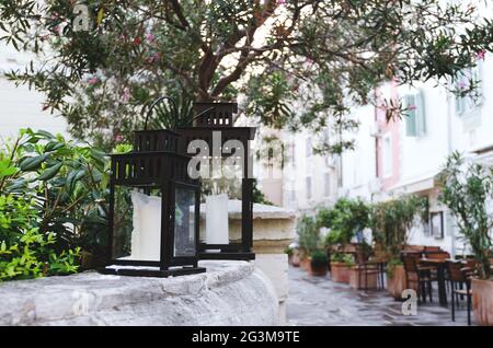 Taschenlampe Kerzenständer, schwarze Vintage dekorative Laternen mit Kerzen steht im Freien in der Stadt, in der Natur mit Blumen, Olivenbaum. Gartengestaltung im Haus Stockfoto