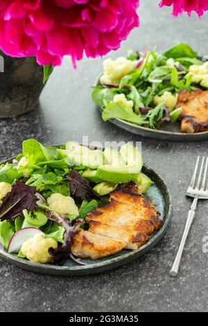 Pfingstrosen in einer Vase und Abendessen auf dem Tisch, ein Teller gebratenes Schweinesteak und ein gemischter Salat mit Blumenkohl und Avocado Stockfoto