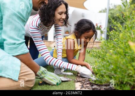 Lächelnde hispanische Mutter, Vater und Tochter im Garten, knien und Pflanzen in Blumenbeet Stockfoto