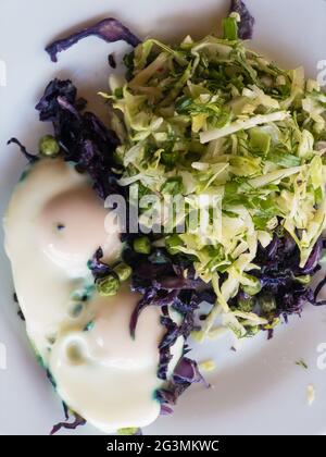Draufsicht auf weißem Teller mit zwei Spiegeleiern, die auf gebratenem Rotkohl mit grüner Erbse liegen und mit frisch gehacktem Kohl garniert werden Stockfoto