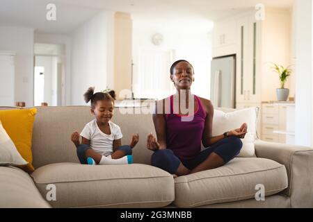 Glückliche afroamerikanische Mutter und Tochter praktizieren Yoga und meditieren auf der Couch im Wohnzimmer Stockfoto