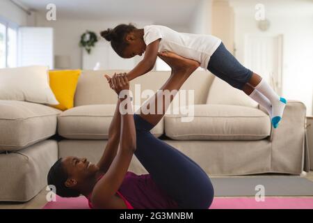 Lächelnde afroamerikanische Mutter, die im Wohnzimmer trainiert, auf dem Rücken liegend und die Tochter auf den Füßen balanciert Stockfoto