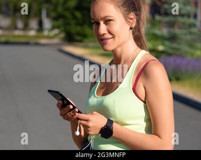 Junge Frau wählt Musik für ihren morgendlichen Lauf. Sie hält ein Smartphone mit Kopfhörern, lächelt und schaut auf die Kamera. Stockfoto