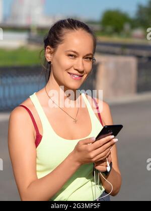 Vertikales Foto einer jungen Frau wählt Musik für ihren morgendlichen Lauf. Sie hält ein Smartphone mit Kopfhörern, lächelt und schaut auf die Kamera. Stockfoto