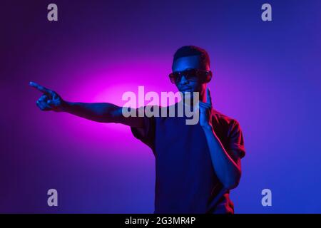 afroamerikanischer junger Mann auf dunklem Studiohintergrund in Neon. Konzept der menschlichen Emotionen, Gesichtsausdruck, Jugend, Verkauf, Werbung. Stockfoto