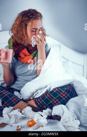 Krankes Mädchen in Schal sitzt auf dem Bett und bläst ihre Nase Stockfoto