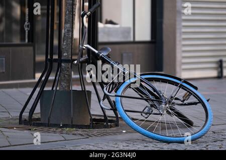 Schwarzes Fahrrad mit blauem Reifen liegt zur Hälfte auf dem Boden und zur Hälfte an einem Baum in einer Einkaufsstraße, unachtsam geparkt oder umgefallen. Gefallenes Fahrrad Stockfoto