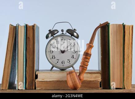 Stapel von alten Büchern, Rauchrohr und Wecker auf dem Holzregal vor dem Hintergrund der weißen Wand Stockfoto