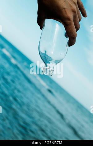Ein Tropfen Wasser fällt aus einer leeren, wiederverwendbaren Glasflasche, kopfüber in der Hand eines jungen kaukasischen Mannes, vor dem Ozean Stockfoto