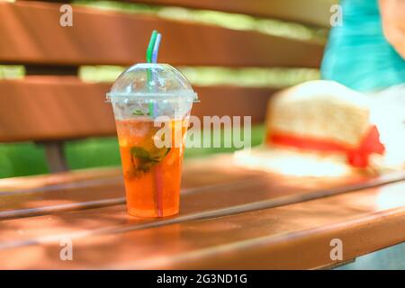 Nahaufnahme eines Plastikbechers zum Mitnehmen mit aperol Spritz Cocktail auf einer Holzbank im Morgengarten. Sonniger Tag. Natur Hintergrund. Stockfoto