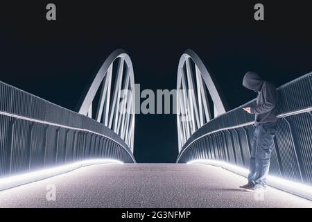 Eine geheimnisvolle, mit einer Kapuze versehene Figur, die auf einem Pfad auf sein Telefon schaut. Silhouetten gegen helle Lichter in der Nacht. Stockfoto