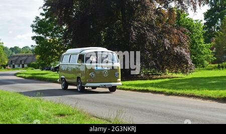 Klassischer Volkswagen Camper Van, der durch Dorfhaus und Bäume im Hintergrund fährt. Stockfoto