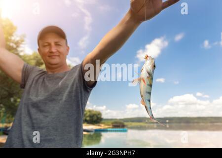 Junge Erwachsene glücklich kaukasischen männlichen Angler Person zeigt frischen Fang von kleinen Barsch Bass Fisch Fishing auf See oder Fluss an hellen warmen sonnigen Tag Stockfoto