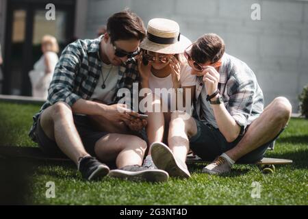 Drei junge Freunde Sitzen auf dem Rasen im Freien und Blick auf Handy. Stockfoto
