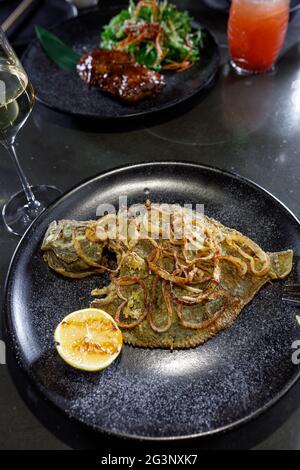 Gebratene Flunder mit Zwiebel und Zitrone auf einem schwarzen Tonteller Stockfoto