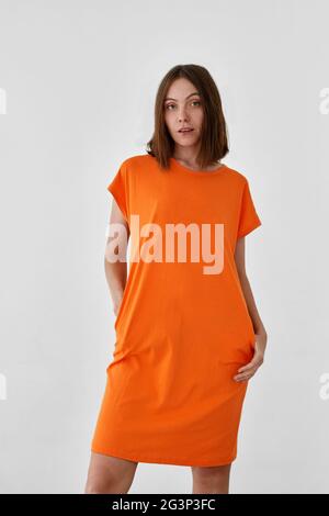Niedliches weibliches Modell in legerem orangefarbenem Kleid mit Blick auf die Kamera vor weißem Hintergrund Stockfoto