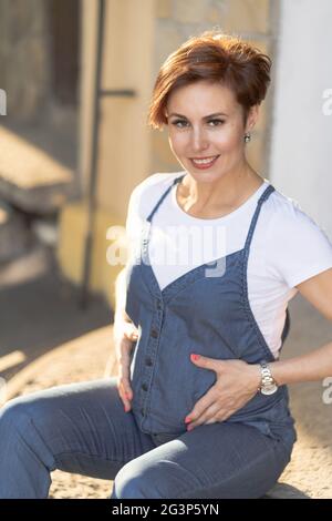 Schwangere Frau in Blau Denim Overalls sitzt auf der Attika Stockfoto