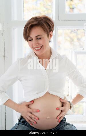 Stilvolle Mumie in weißem Hemd und blauer Jeans sitzt am Fenster. Schwangerschaft Konzept. Stockfoto