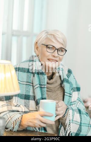 Eine ältere Frau, eingewickelt in EINEN karierten Kariert, trinkt zu Hause Tee Stockfoto