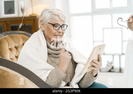 Elegante Ältere Frau In Wollpullover Liest Etwas Angenehmes Auf Ihrem Tablet Stockfoto