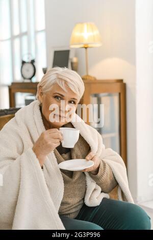 Fröhliche ältere Frau in EINEM warmen Strickmuster eingewickelt entspannend zu Hause Stockfoto